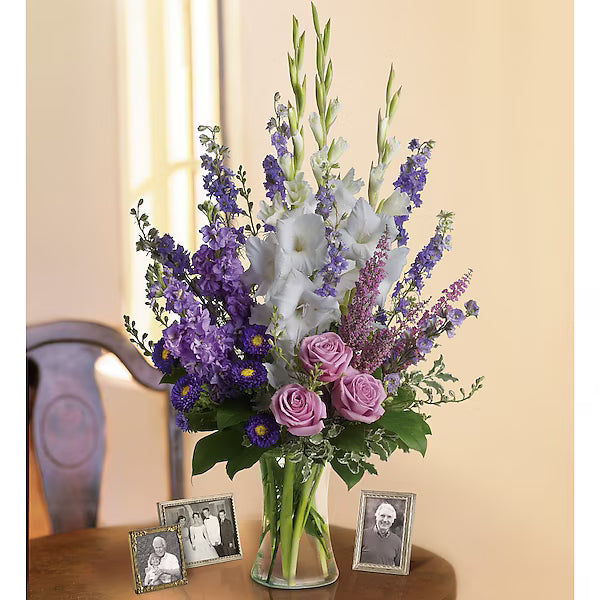 Bouquet of purple, white, and pink flowers in a clear vase on a wooden table with framed photos.